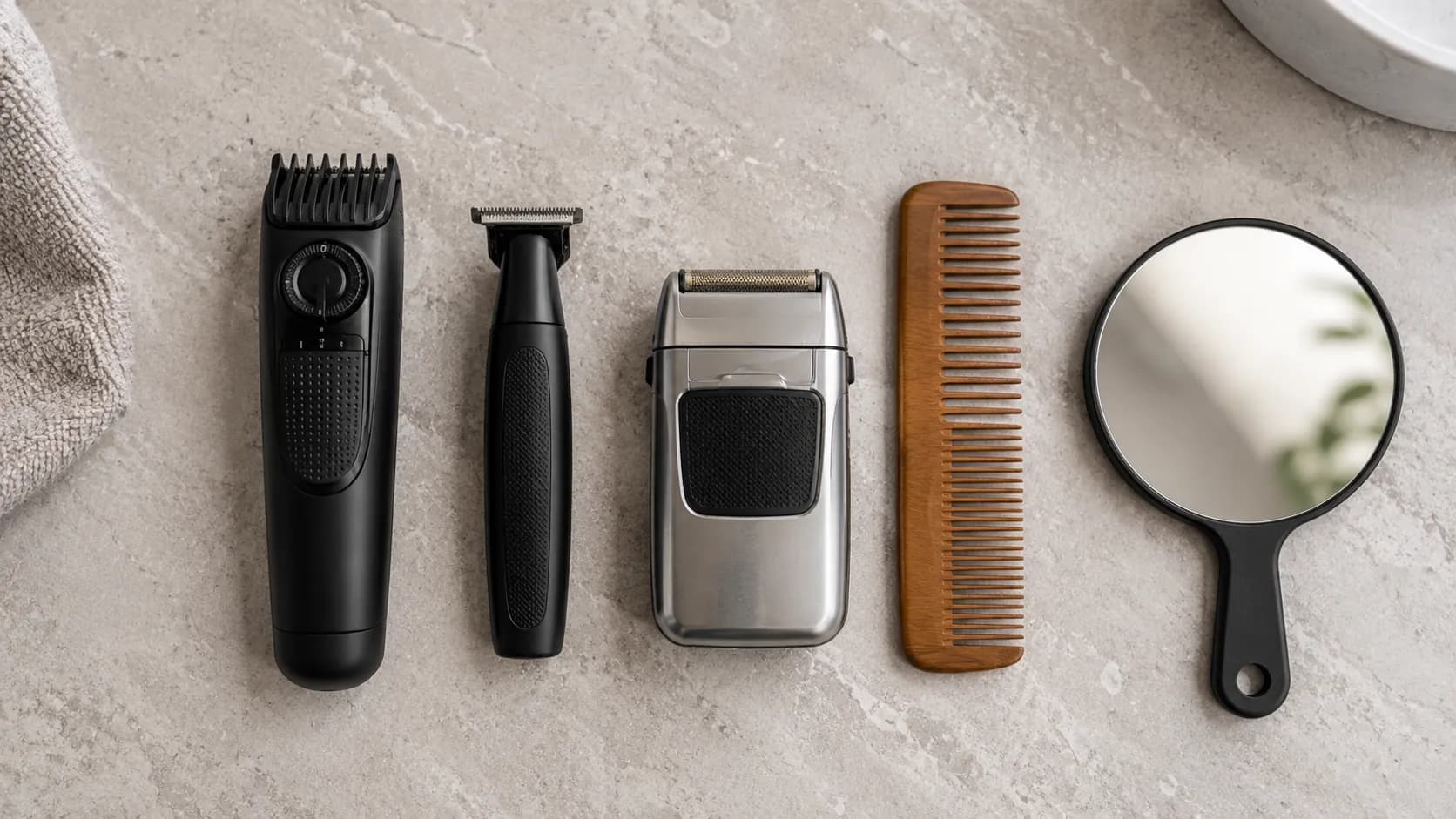 Overhead flat-lay of a beard grooming kit with an adjustable trimmer, T-outliner, foil shaver, wooden comb, and hand mirror arranged neatly on a light stone bathroom countertop.